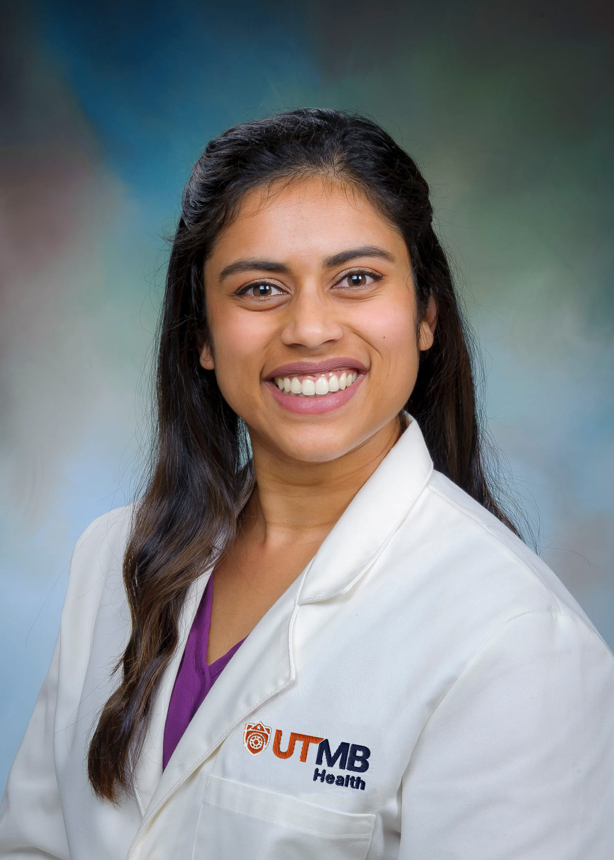 Dr. Radhini Bell Portrait of a person wearing a white UTMB Health lab coat, facing the camera against a blue-green studio background.