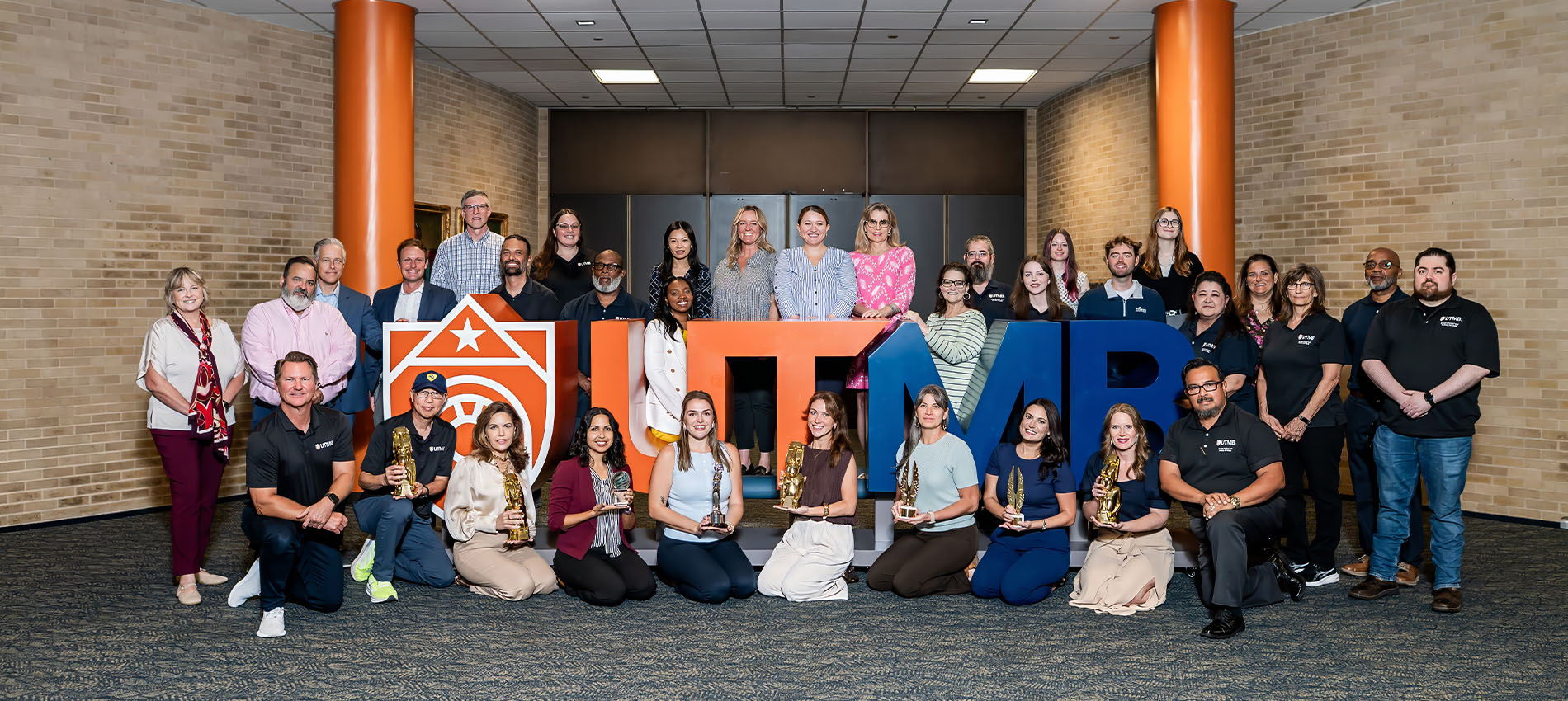 A group of people posed indoors around large “UTMB” letters and a shield-shaped sign, with several individuals holding award trophies.