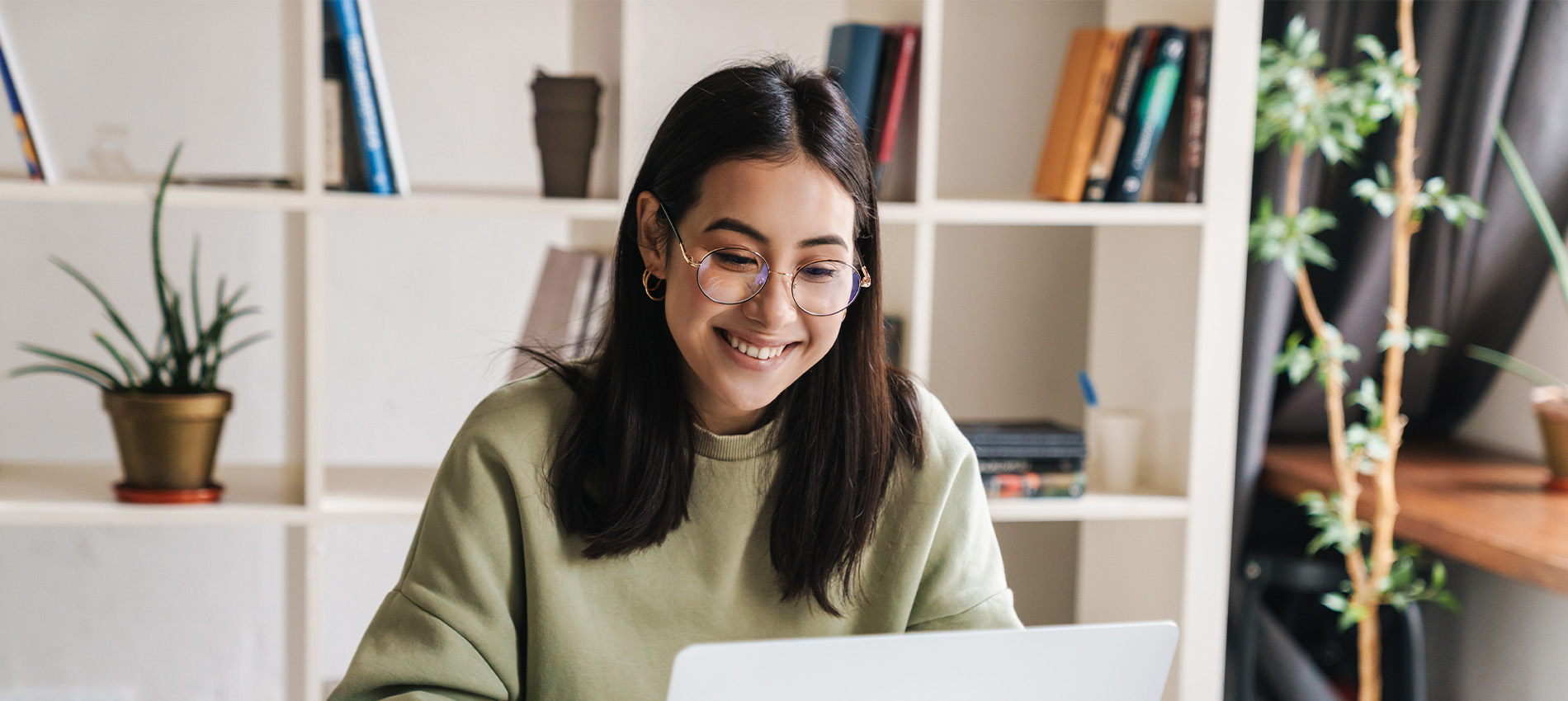 Person working at a laptop in a home office with shelves and plants in the background.