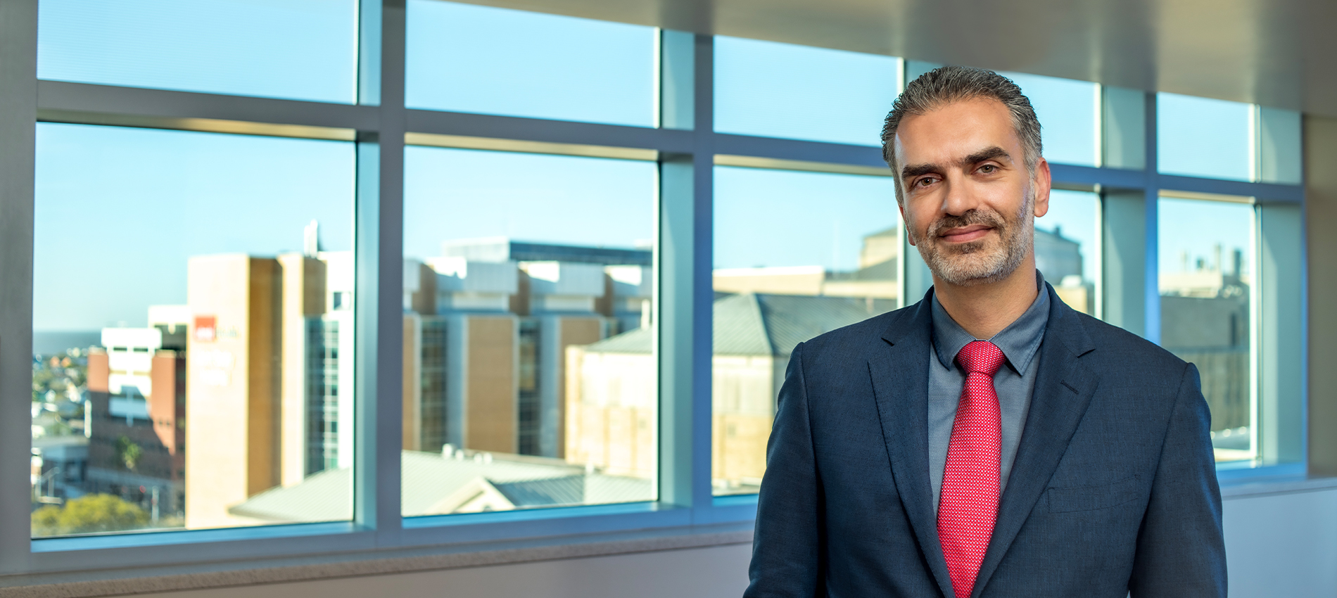 Person in a suit and red tie standing indoors with large windows showing a hospital in the background.