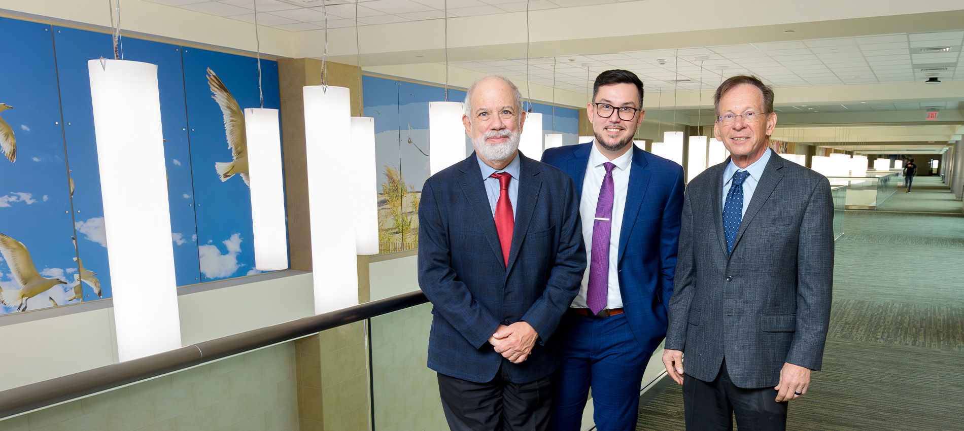 Three people in business suits stand side by side in a brightly lit indoor hallway with large hanging light fixtures. A wall featuring bird illustrations is visible behind them.