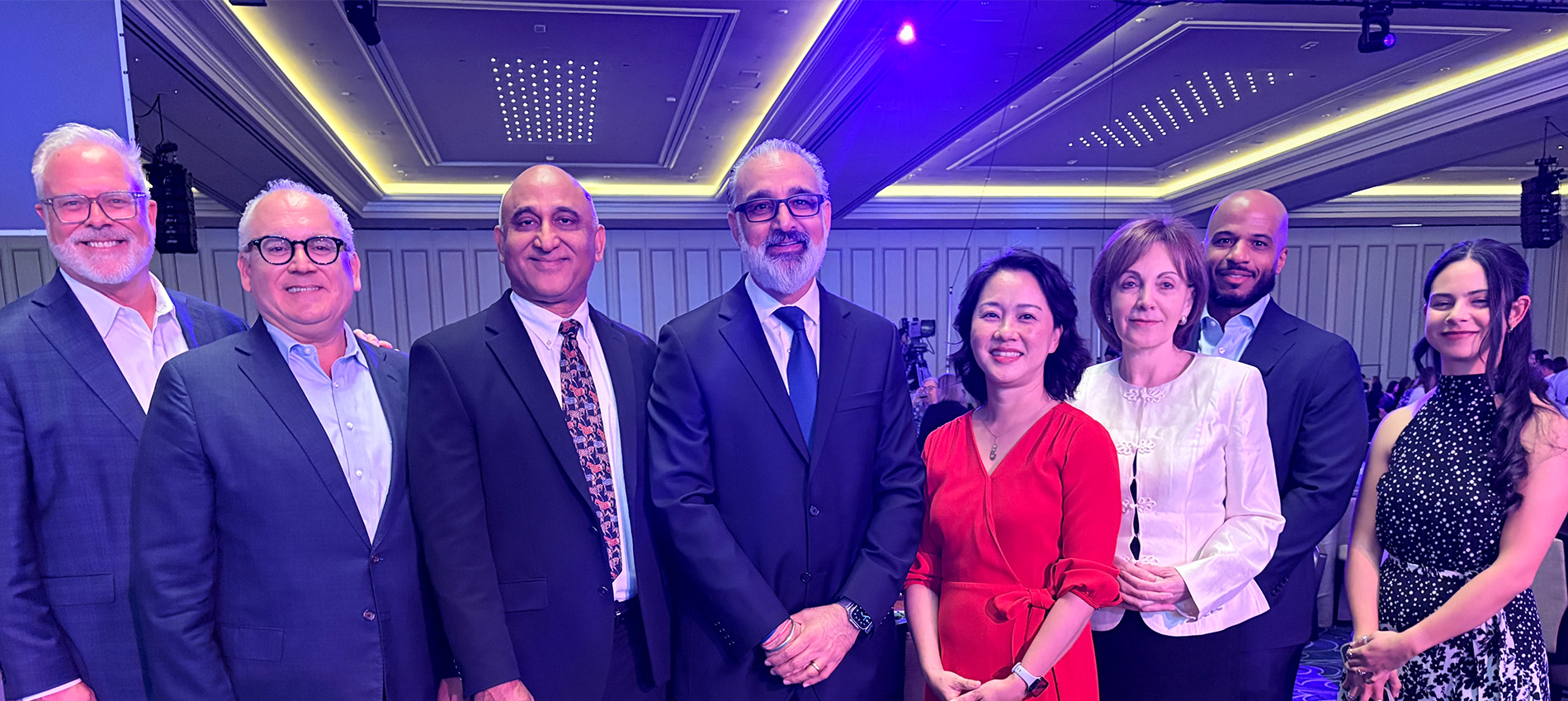 Seven UTMB Health leaders and representatives pose for a group photo in a ballroom during the 2026 Vizient Connections Summit in Las Vegas, with banquet tables, attendees, and purple event lighting visible in the background.