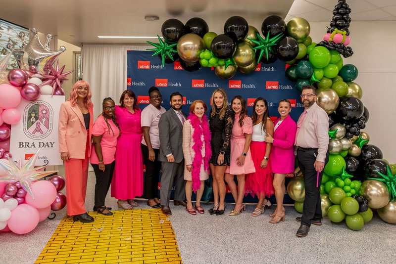 BRA Day 2025 Group of people posing in front of a UTMB Health backdrop framed by pink, green, black, and gold balloon displays, with a BRA Day sign visible on the left.