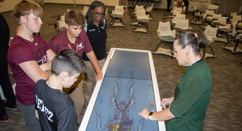 A group of students and an adult gather around a large interactive display showing a detailed anatomical vascular model.