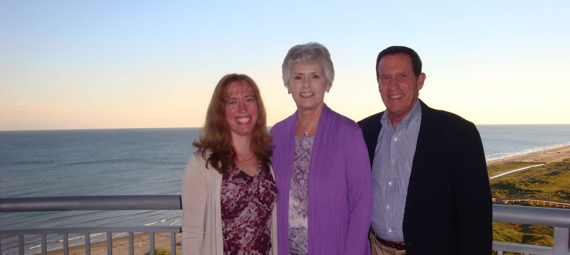 A retired couple and their adult daughter wearing dressy attire pose on a balcony in front of a beach at sunset.