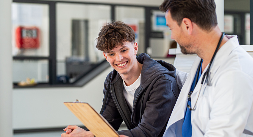 Clinician in a white coat with a stethoscope reviews a clipboard while seated beside a patient in a clinical exam area with glass-walled rooms in the background.