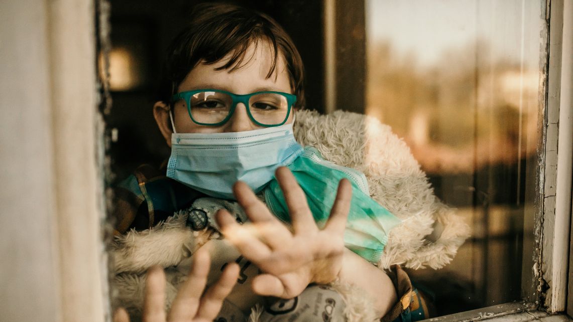 A young boy in green glasses and surgical mask looks out a window.