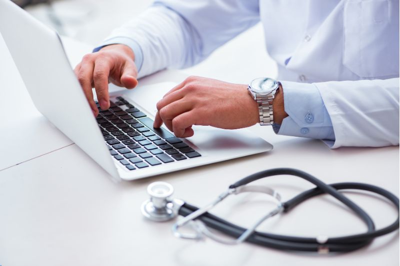 A doctor in a white coat works on a laptop with his stethoscope next to him