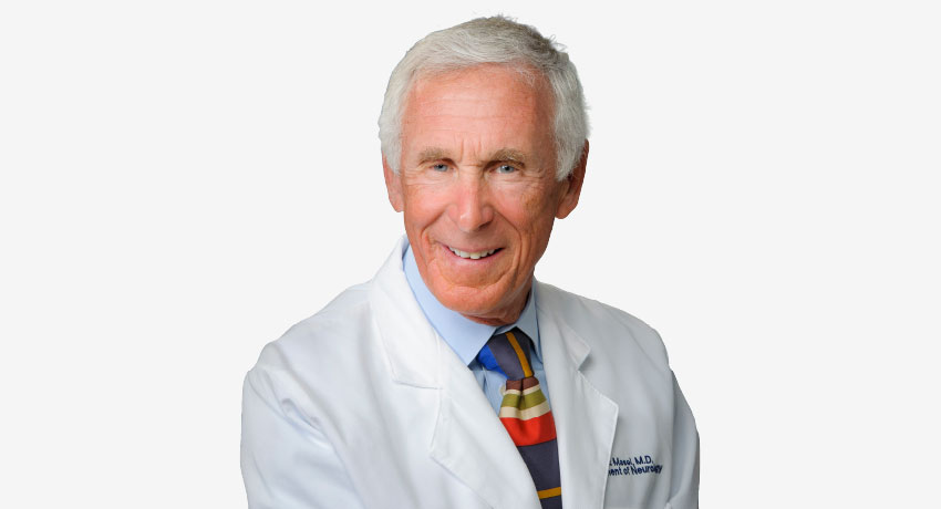 A person wearing a white medical coat and a multicolored striped tie stands against a plain background. The coat includes embroidered text on the left side.