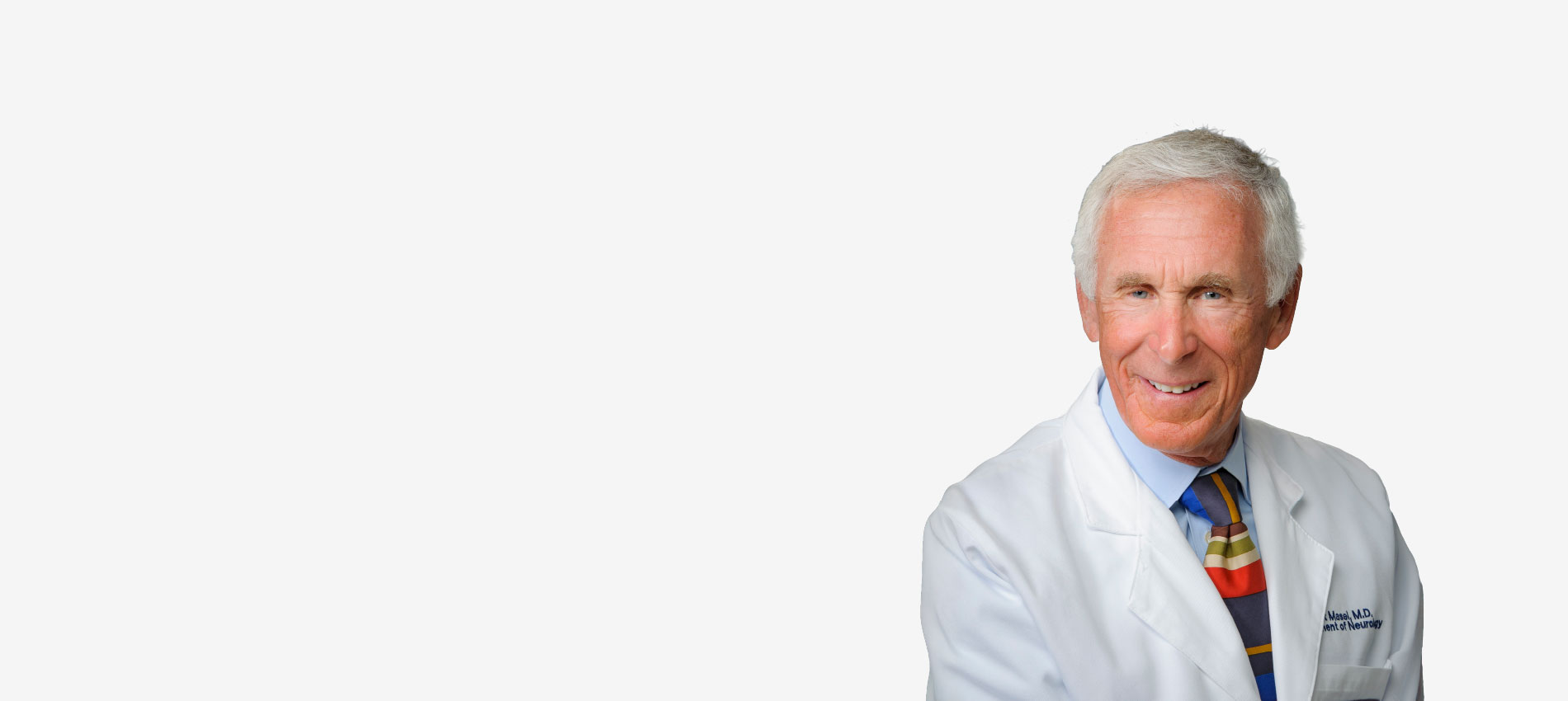 A person wearing a white medical coat and a multicolored striped tie stands against a plain background. The coat includes embroidered text on the left side.