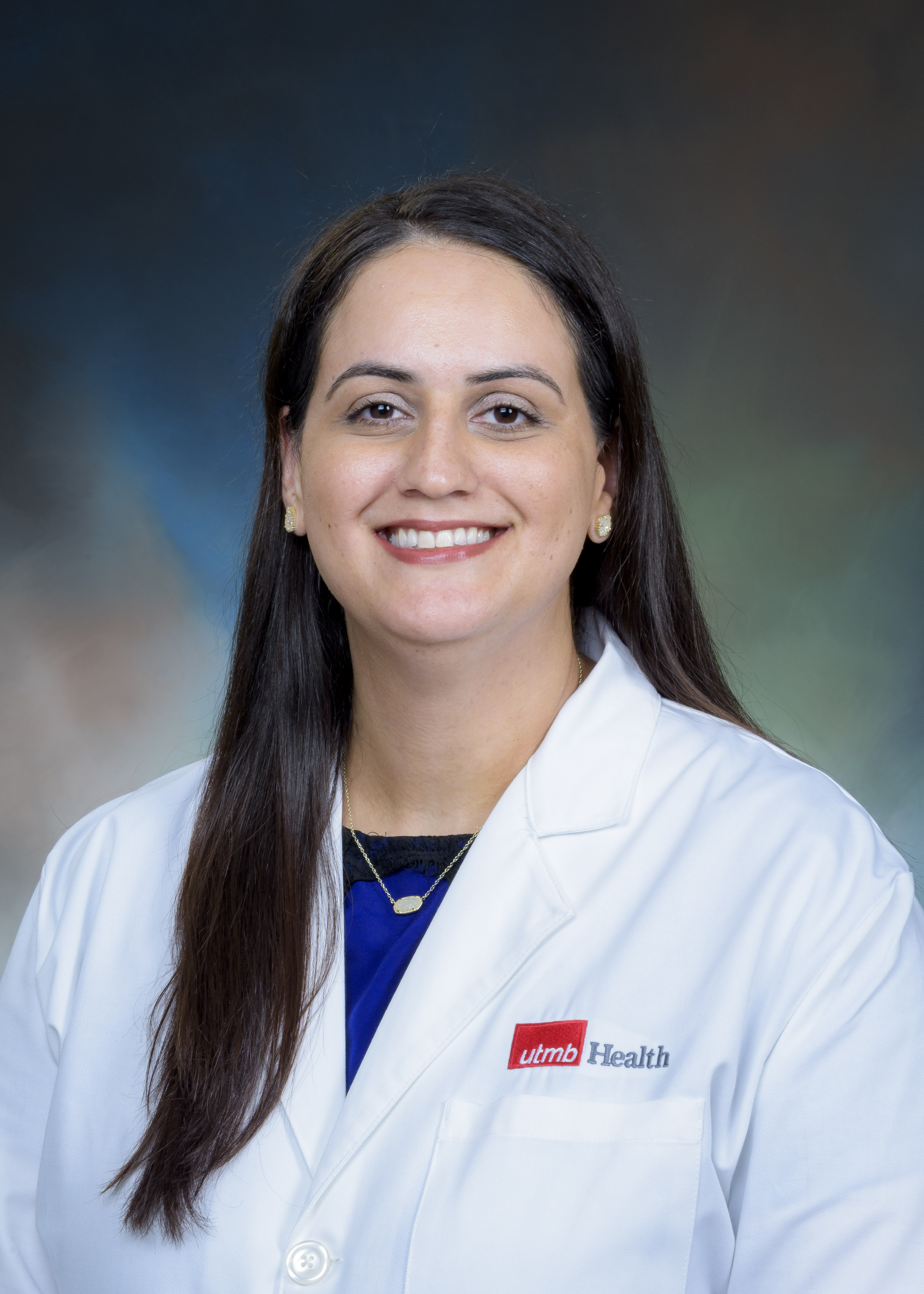 Person wearing a white lab coat with a UTMB Health logo over a blue top, posed against a neutral studio background. The face is intentionally blurred.