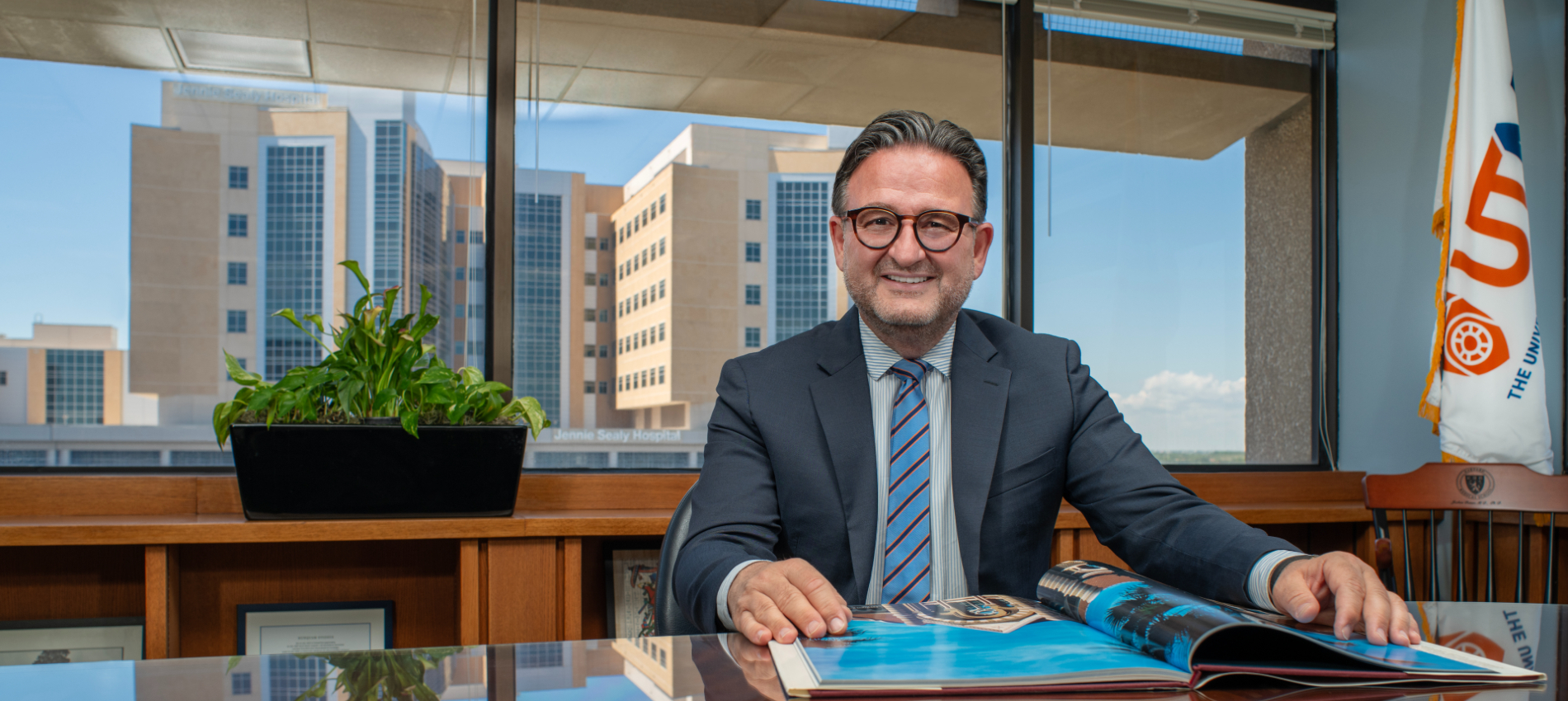 A man in a suit with short dark hair and black glasses stands indoors by a window holding books, with the Jennie Sealy Hospital building visible outside.