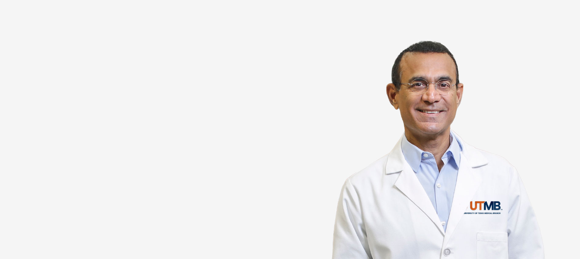 Portrait of a person wearing a white UTMB lab coat over a light blue collared shirt, standing against a plain light gray background. The UTMB logo is visible on the lab coat.