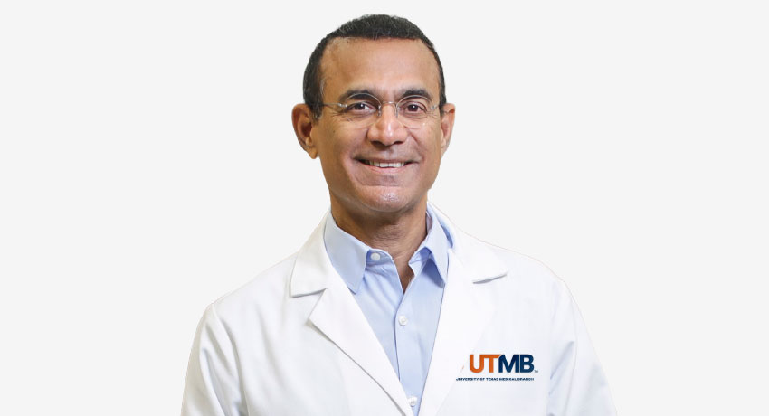 Portrait of a person wearing a white UTMB lab coat over a light blue collared shirt, standing against a plain light gray background. The UTMB logo is visible on the lab coat.