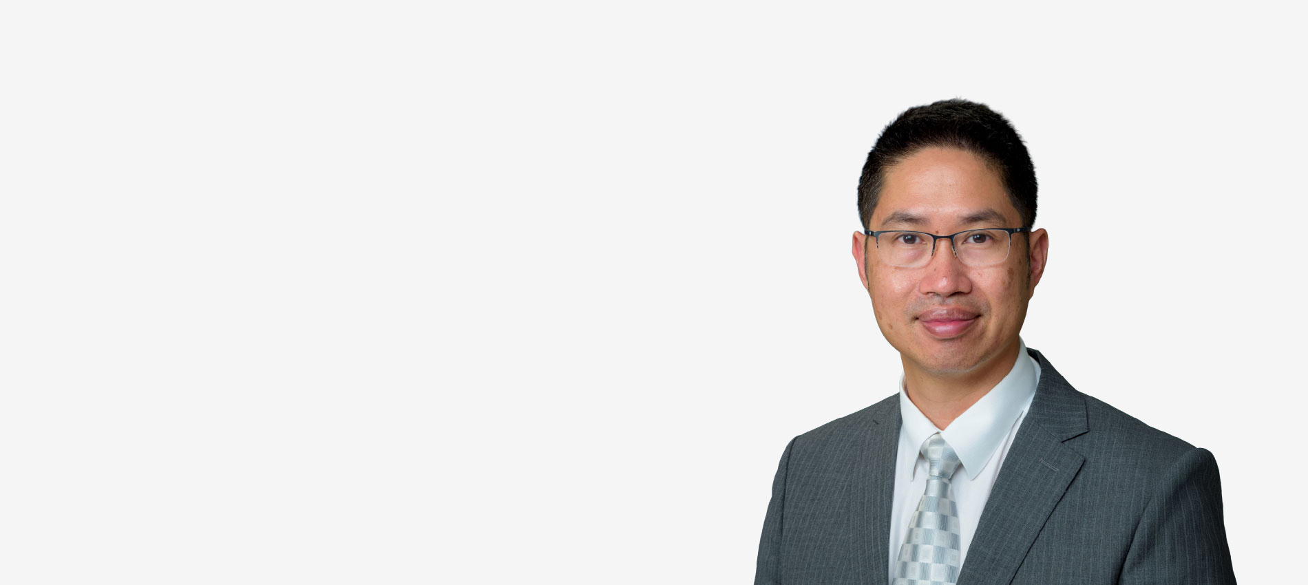 Portrait shows a man wearing glasses, a gray suit jacket, white dress shirt and patterned tie, facing the camera against a light gray background