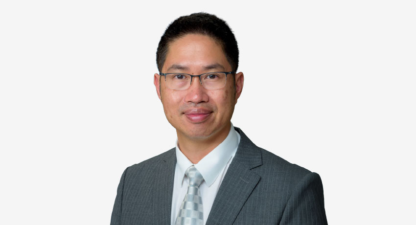 Portrait shows a man wearing glasses, a gray suit jacket, white dress shirt and patterned tie, facing the camera against a light gray background