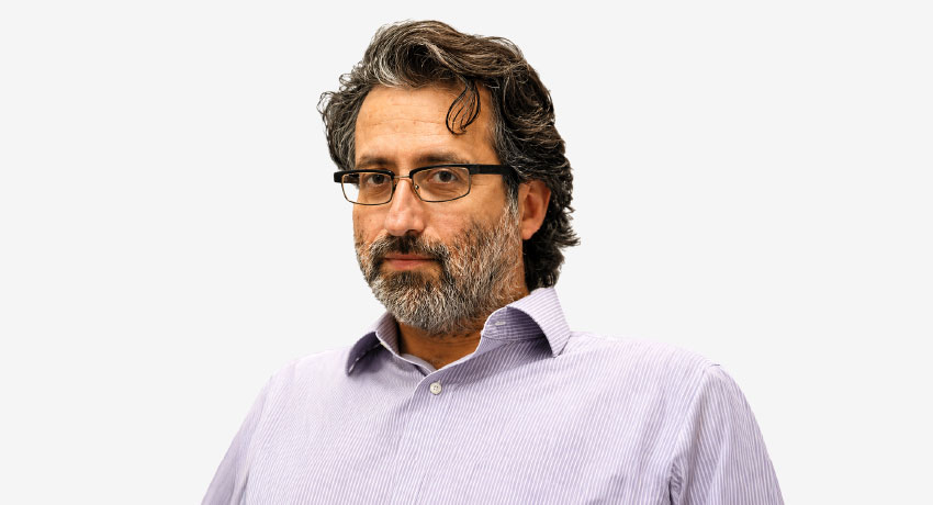 Middle-aged man with salt-and-pepper hair and a full beard, wearing rectangular glasses and a light purple striped button-down shirt, looking directly at the camera with a neutral expression against a plain light gray background.