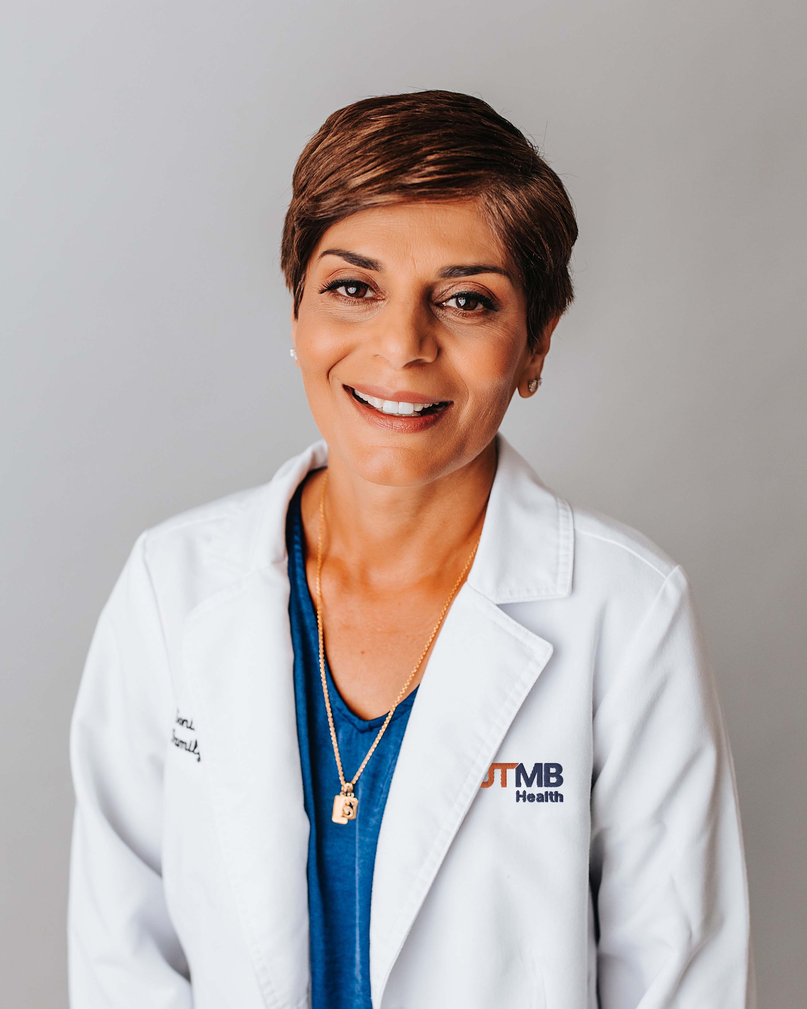 Upper-body view of a clinician wearing a white lab coat with the UTMB Health logo over a blue top, standing against a neutral background