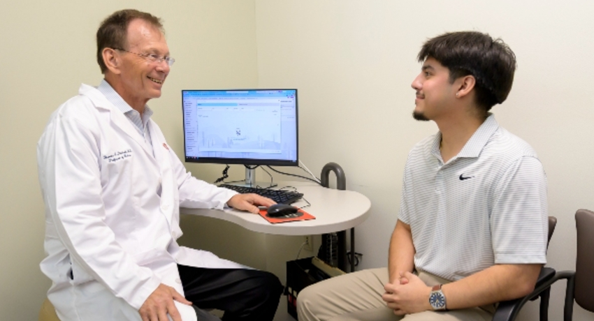 A doctor in a white coat sits in an exam room speaking with a young man seated across from him beside a computer workstation. Both are smiling and engaged in conversation during a medical appointment.