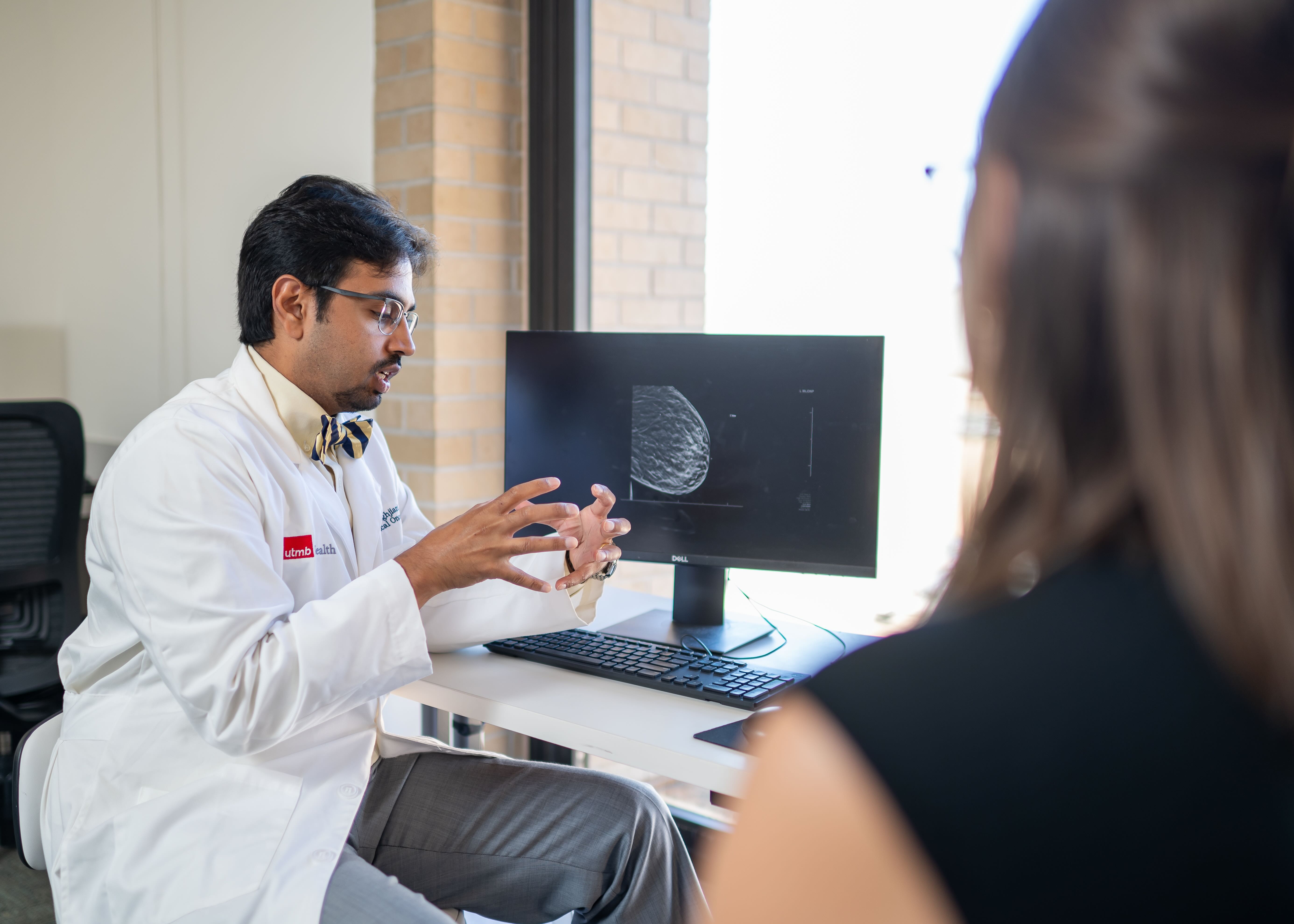 A health care provider in a white coat sits at a desk and gestures toward a computer monitor displaying breast imaging while speaking with a patient during an office visit