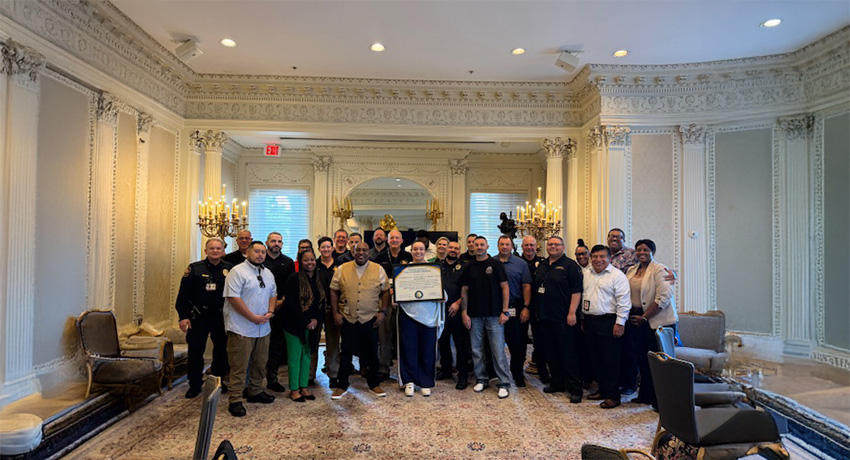 Group of UTMB Police Department personnel standing together in an ornate meeting room while a team member at the center displays a framed dual accreditation certificate recognizing achievement through CALEA and IACLEA.