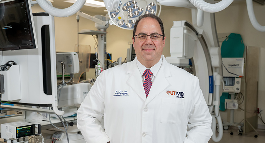 Clinician wearing a white lab coat with the UTMB Health logo stands in a cardiac procedure room with imaging equipment and monitors visible behind them.