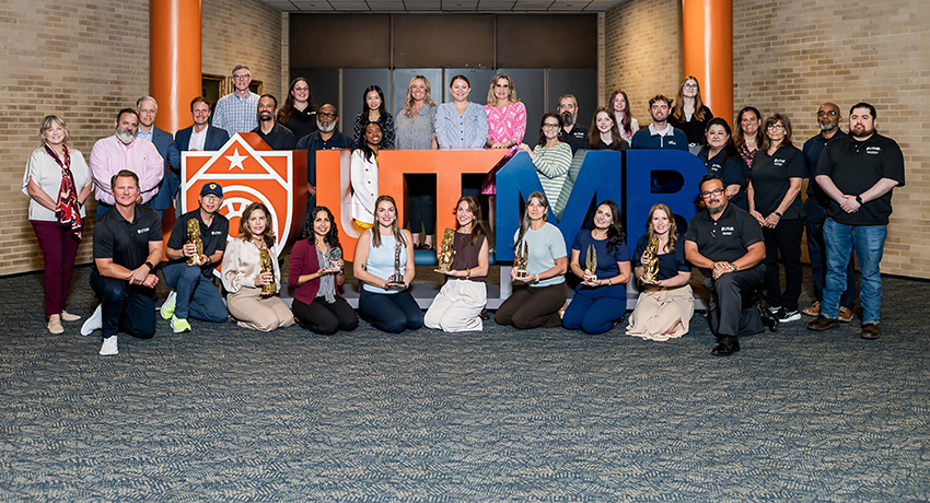 A group of people is posed indoors around large “UTMB” letters and a shield-shaped sign, with several individuals holding award trophies.