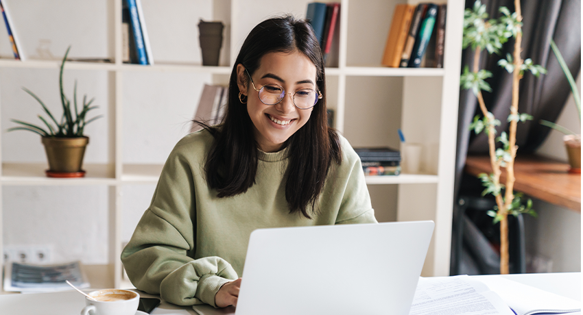 Person working at a laptop in a home office with shelves and plants in the background.