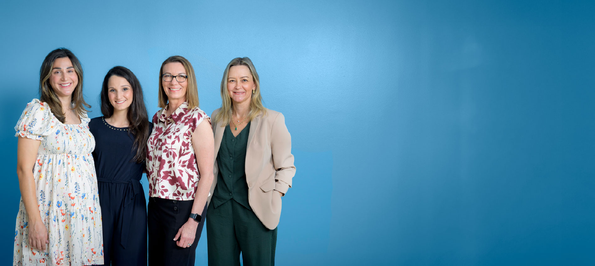 Four women in business casual attire stand close together in front of a blue background.