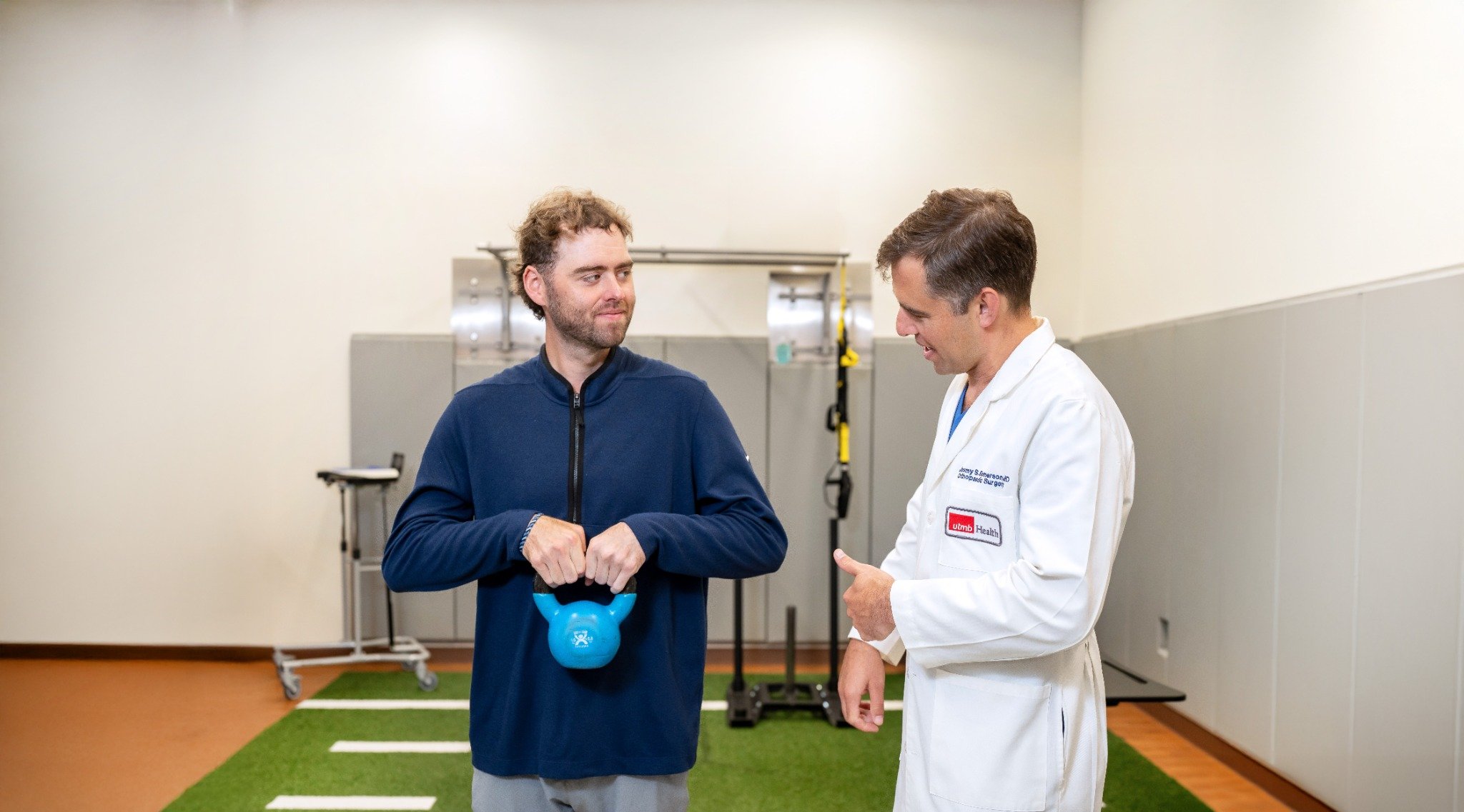 Health care provider in a white coat stands beside a patient holding a blue kettlebell in a clinical gym, with exercise equipment and turf flooring visible, illustrating guided physical activity and movement support in a medical setting