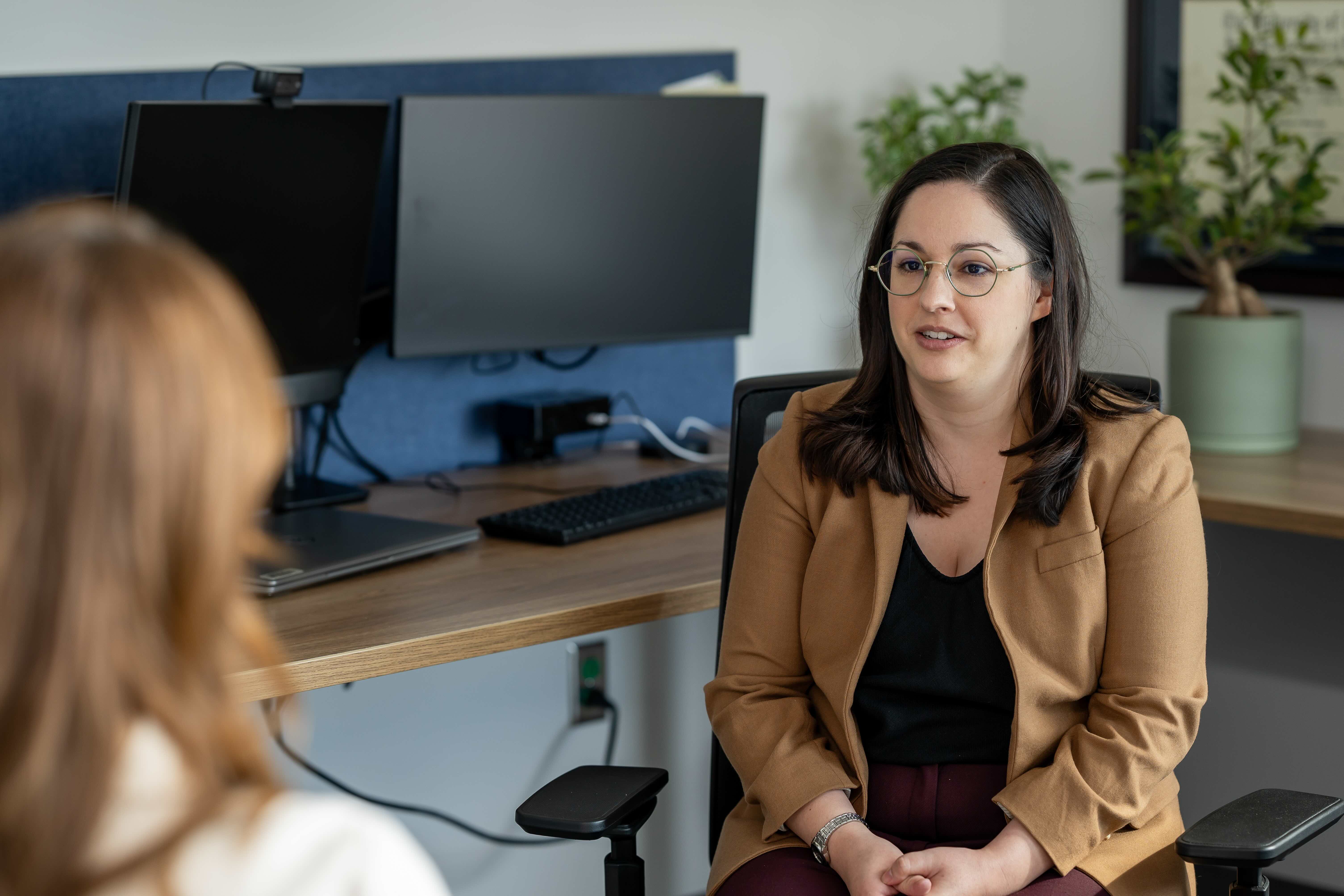 A health care provider wearing a tan jacket sits facing a patient during an office consultation, with computer monitors, a desk and a plant visible in the background