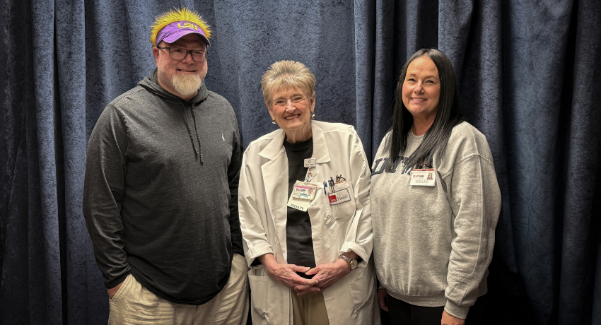 Three people stand together smiling in front of a dark curtain backdrop. The person in the center wears a white lab coat with name badges, while the two on either side are dressed casually, one in a hoodie and cap and the other in a sweatshirt.