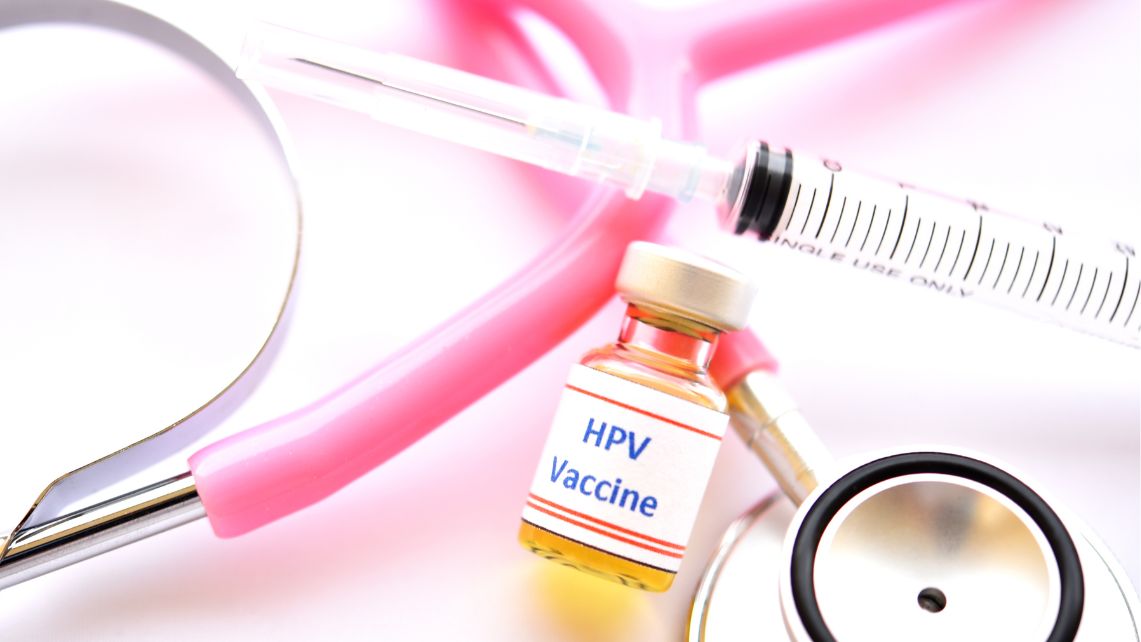A vial labeled HPV vaccine and syringe laying across a pink stethoscope