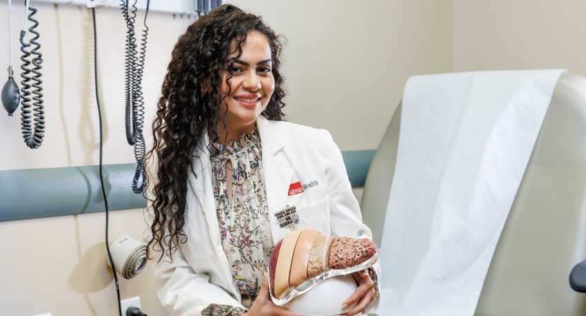 Dr. Randa Abdelmasih smiles while seated in a medical exam room, holding an anatomical model of a fatty liver on her lap. An exam table and medical equipment are visible behind her