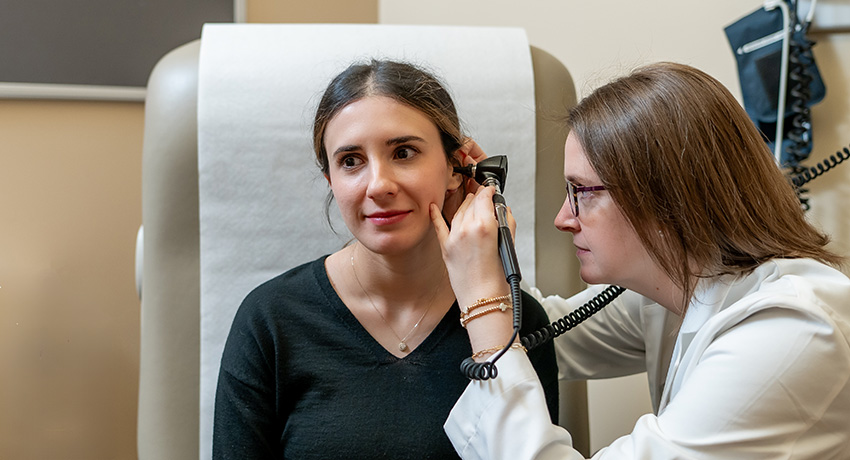 Clinician uses a handheld otoscope to examine a seated patient’s ear in a clinical exam room.