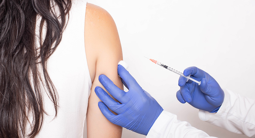 A clinician wearing blue gloves prepares to give an injection into a person’s upper arm, holding a cotton pad against the skin.