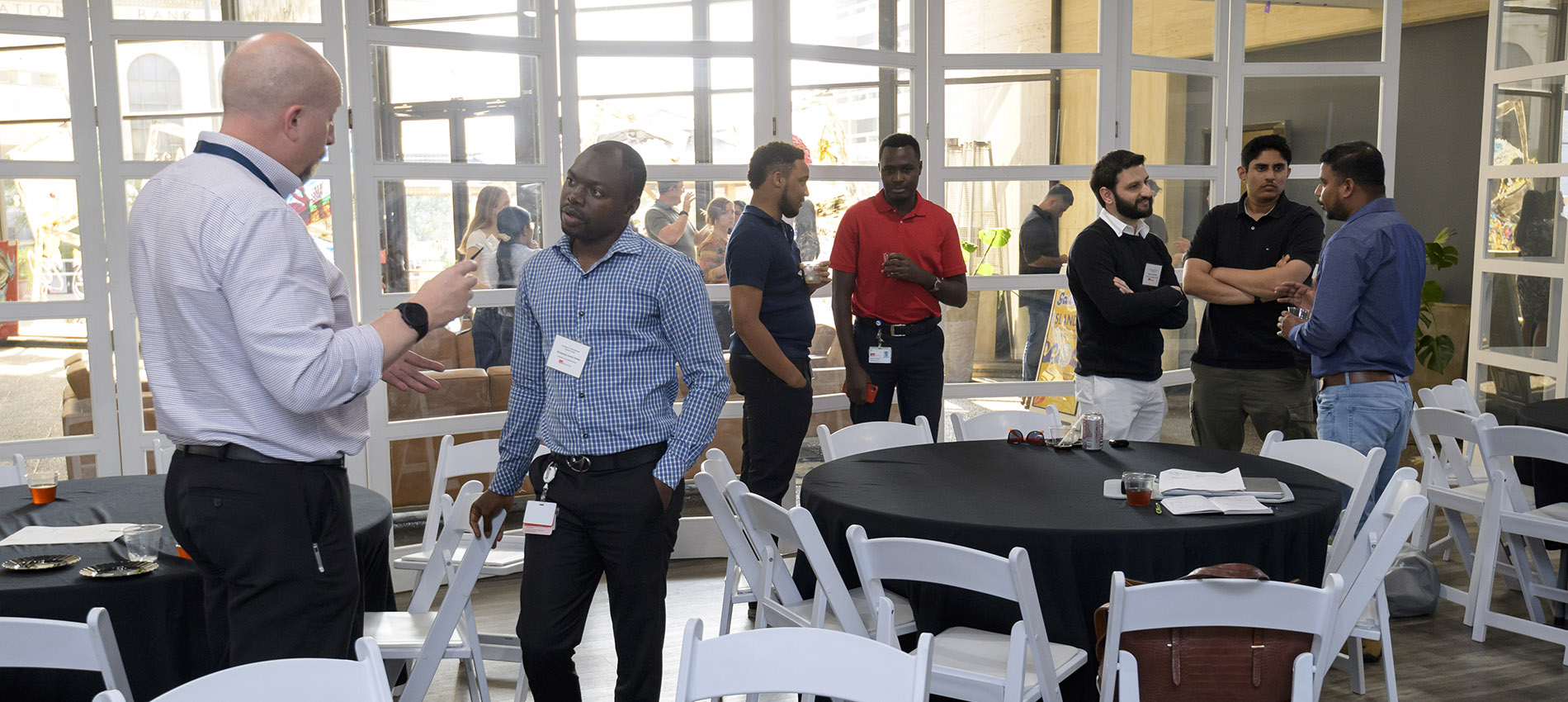Participants in the 2025 Innovation Summer camp stand and chat in three different small groups in a well-lit room with tables and chairs nearby.