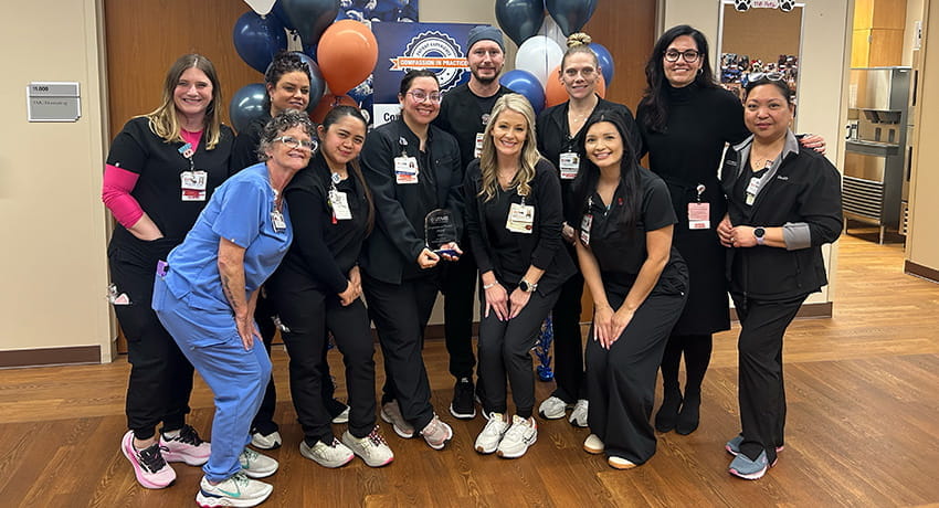 Group of UTMB health care staff standing together indoors, posing with an award in front of blue and tan balloons.