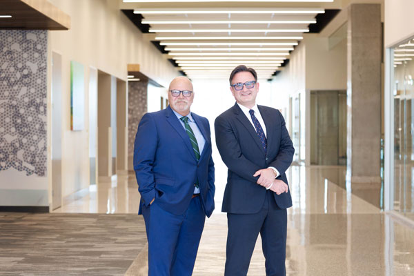 Dr. Giulio Taglialatela (left) with Dr. Jochen Reiser (right). Two people in business suits stand side by side in a bright, modern hallway with reflective flooring and overhead linear lighting.