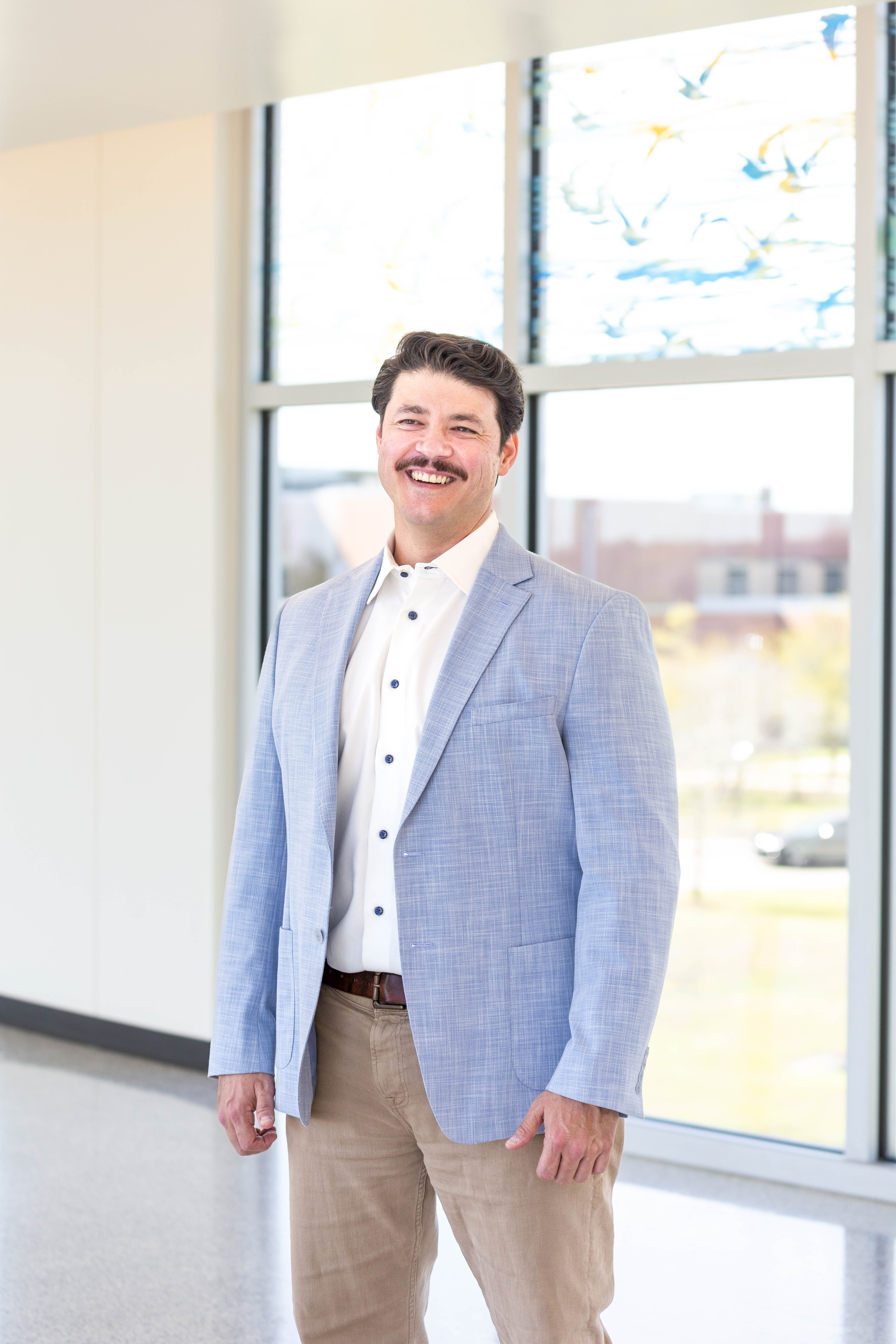 Dr. Joseph Ray, Division Director of Medical Genetics and Metabolism at UTMB. Adult wearing a light jacket stands indoors near large windows with natural light, with an outdoor scene faintly visible through the glass.