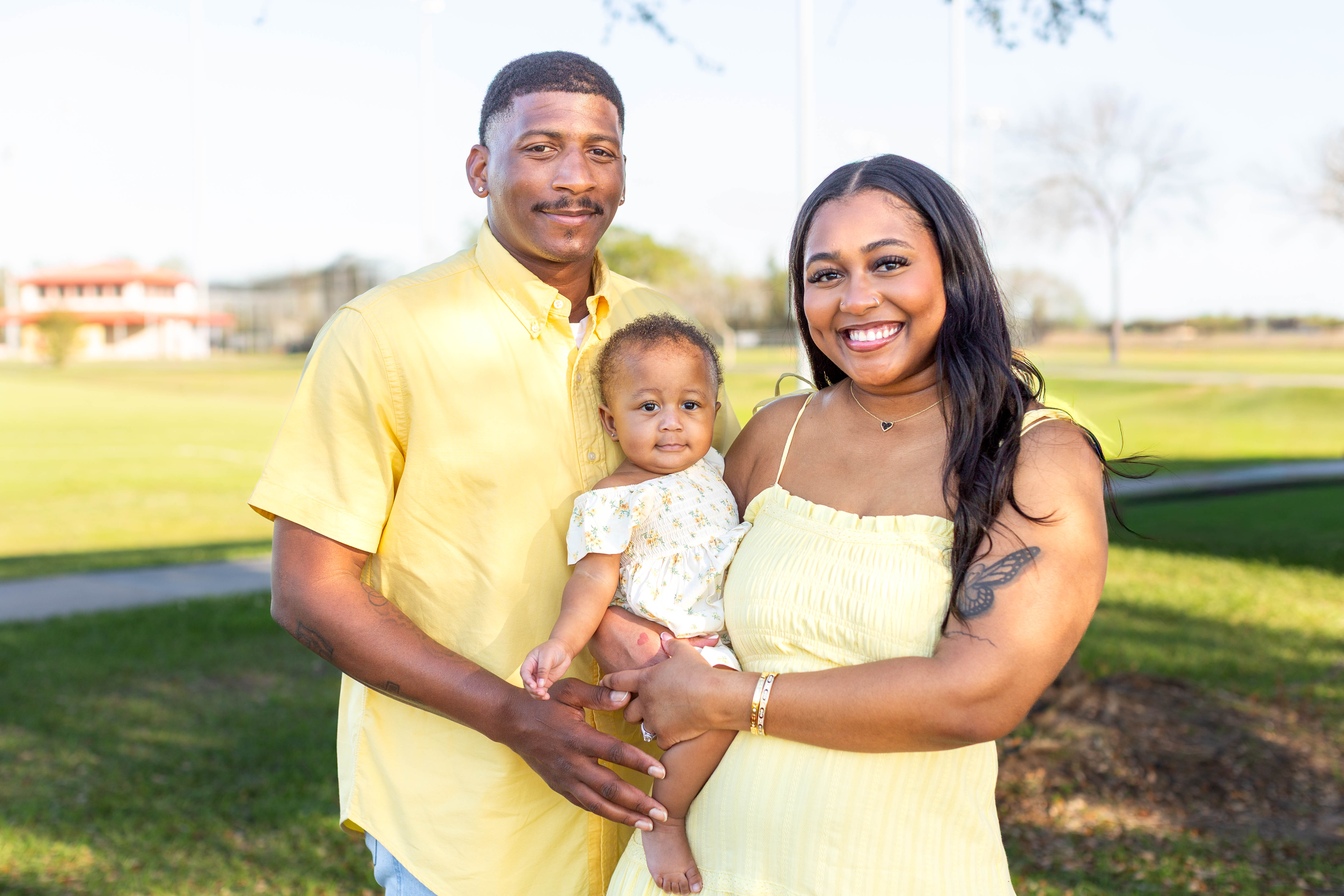 Knova with parents Donta Harris and Sydney Norman. Adult holds an infant while standing beside another adult outdoors, with a grassy area, trees, and a building visible in the background.