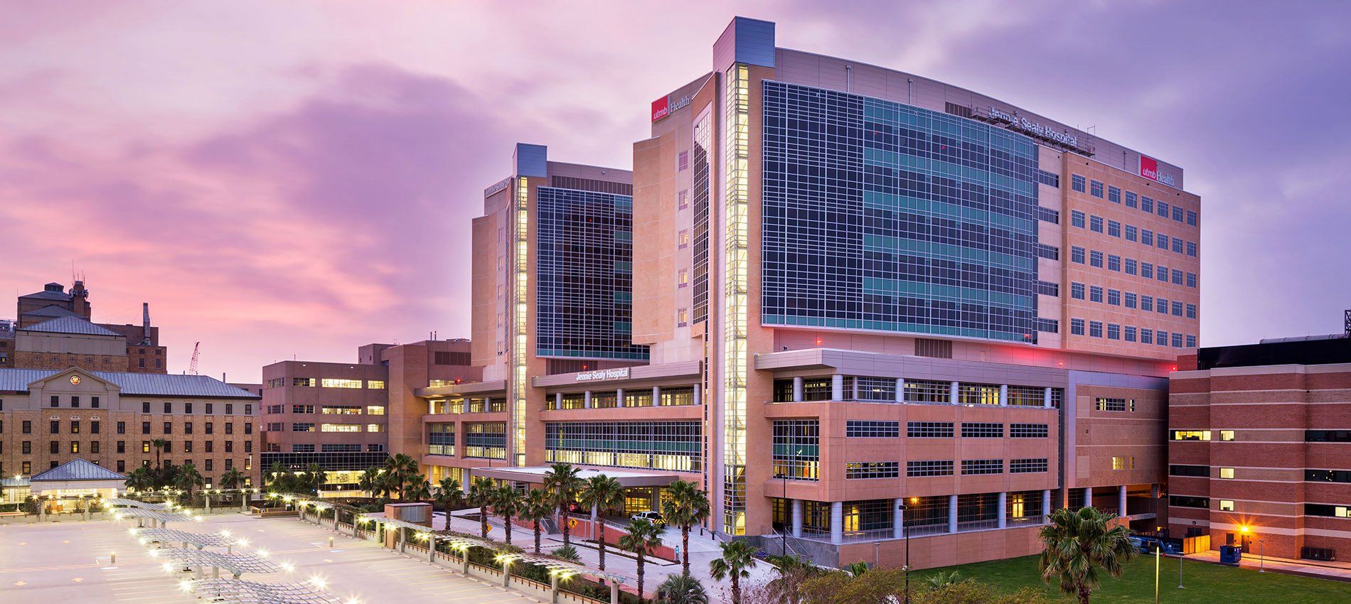 A photography of Jennie Sealy Hospital with interior lights glowing, set against a sunset with pink and purple hues.