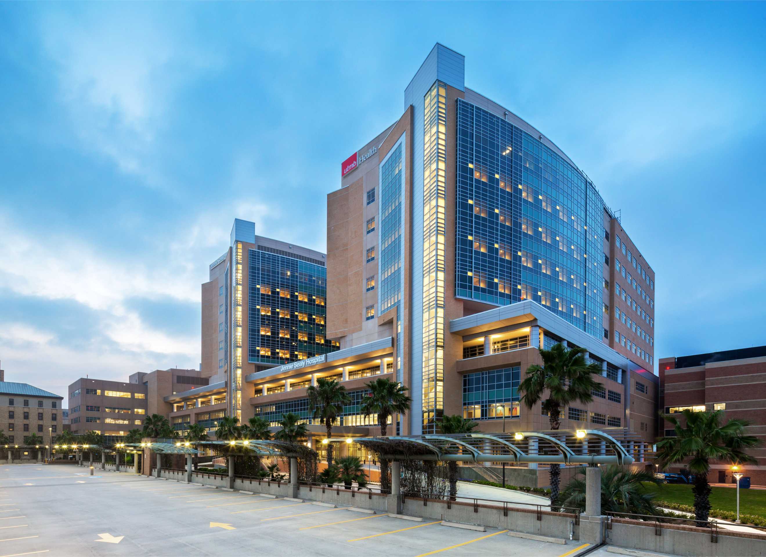 Exterior view of Jennie Sealy Hospital at UTMB, a multistory modern hospital building with glass façades and palm trees, viewed at dusk