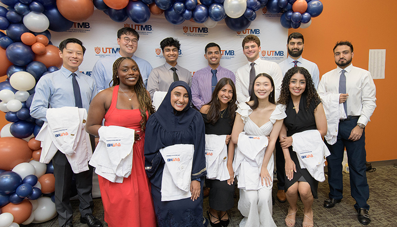 A group of incoming John Sealy School of Medicine students pose beneath a UTMB balloon arch, holding their white coats before the White Coat Ceremony. The students stand in front of a UTMB-branded backdrop as they begin their medical education journey.