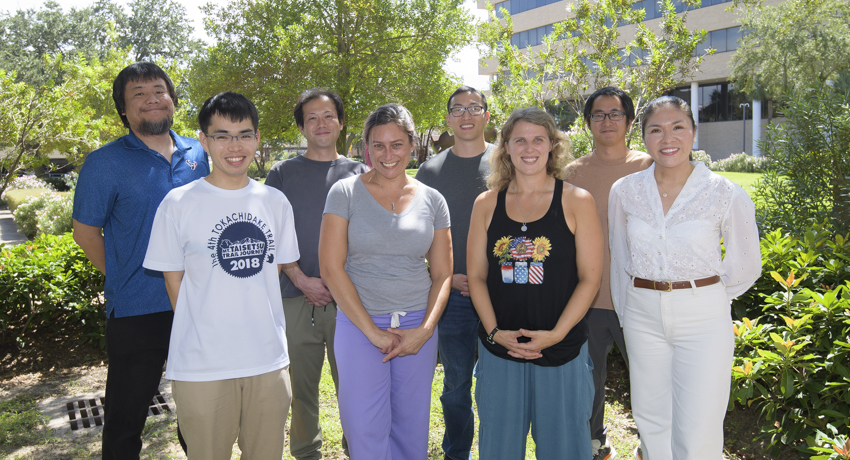 Eight men an women in casual dress pose outdoors on the UTMB campus, surrounded by lush vegetation.
