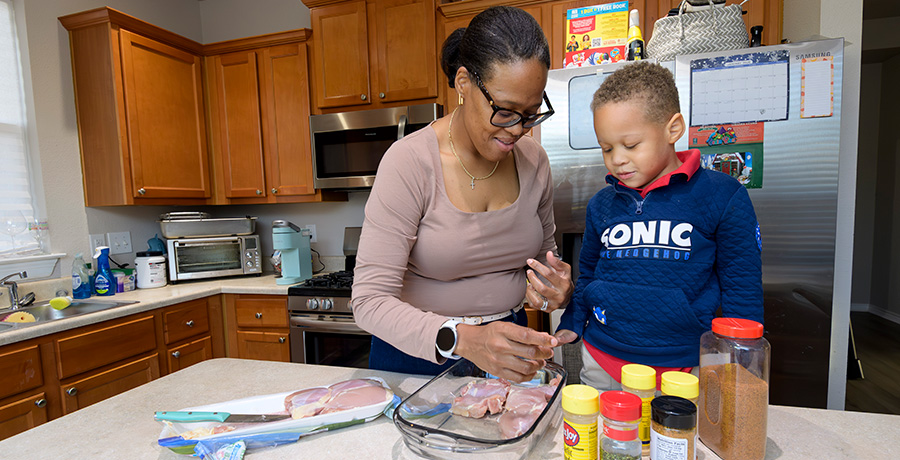 A preschooler standing at the kitchen island with his mother watches her season the chicken.