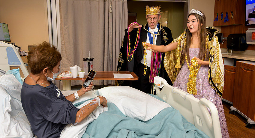 Hospital patient in bed receives Mardi Gras beads from costumed visitors wearing crowns and formal attire inside a patient room.