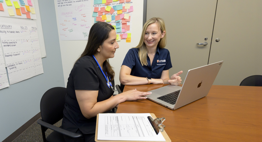 Two women sit at a conference table looking at a laptop during a collaborative meeting in an office. A clipboard with paperwork rests on the table, while colorful sticky notes and planning boards cover the wall behind them.