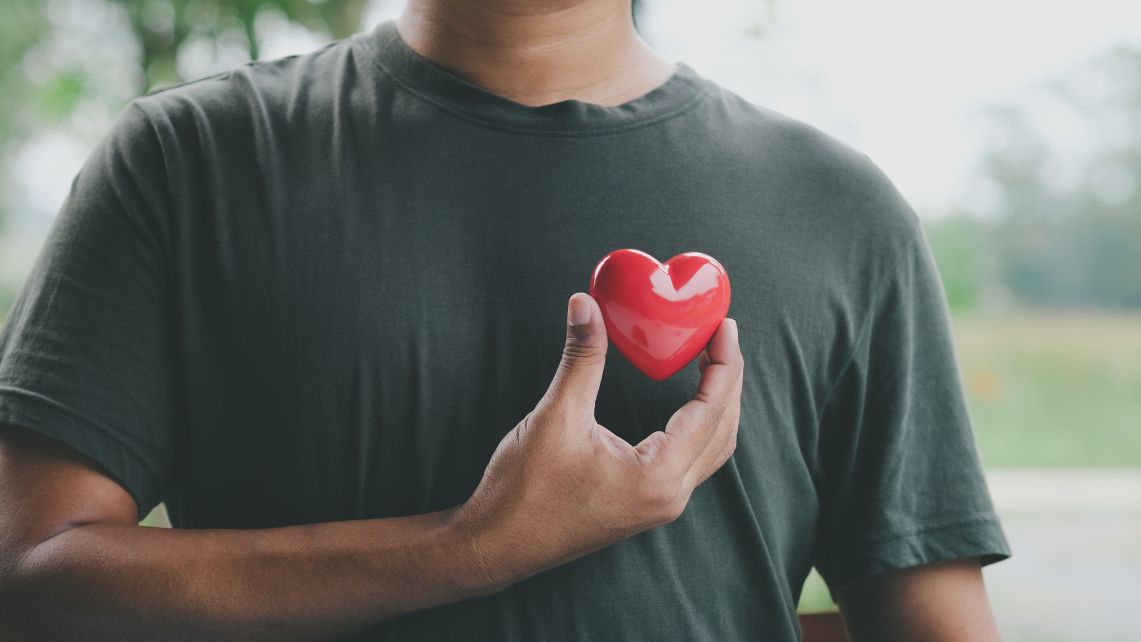 Person wearing a dark shirt holds a small red heart shape against the chest, symbolizing heart health and cardiovascular well‑being.
