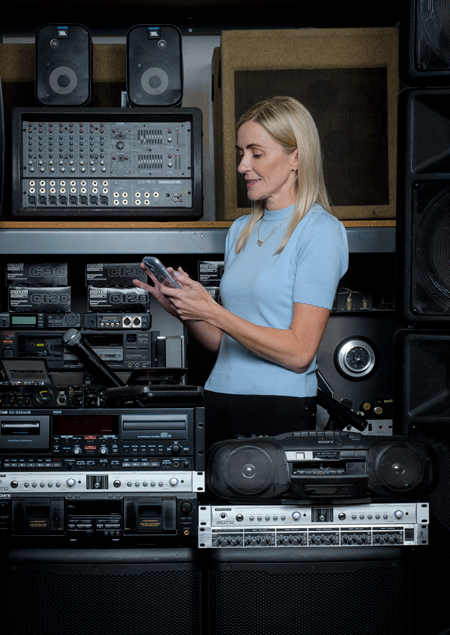 Meredith C. Masel, PhD, director of the Oliver Center Person standing among shelves of audio equipment and speakers, looking at a smartphone. Vintage stereo components, cassette decks, and a boombox are visible around the person.